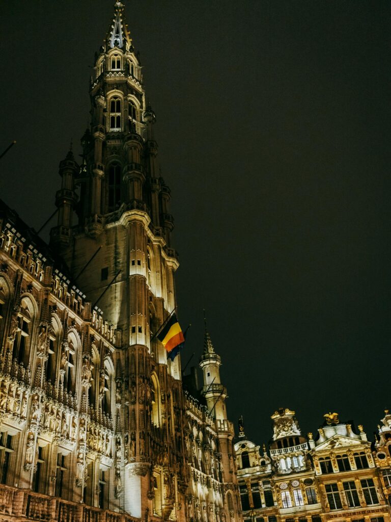 Brussels Grand Place at night: floodlit gothic Hotel de Ville and surrounding guildhalls with Belgian flag, Belgium
