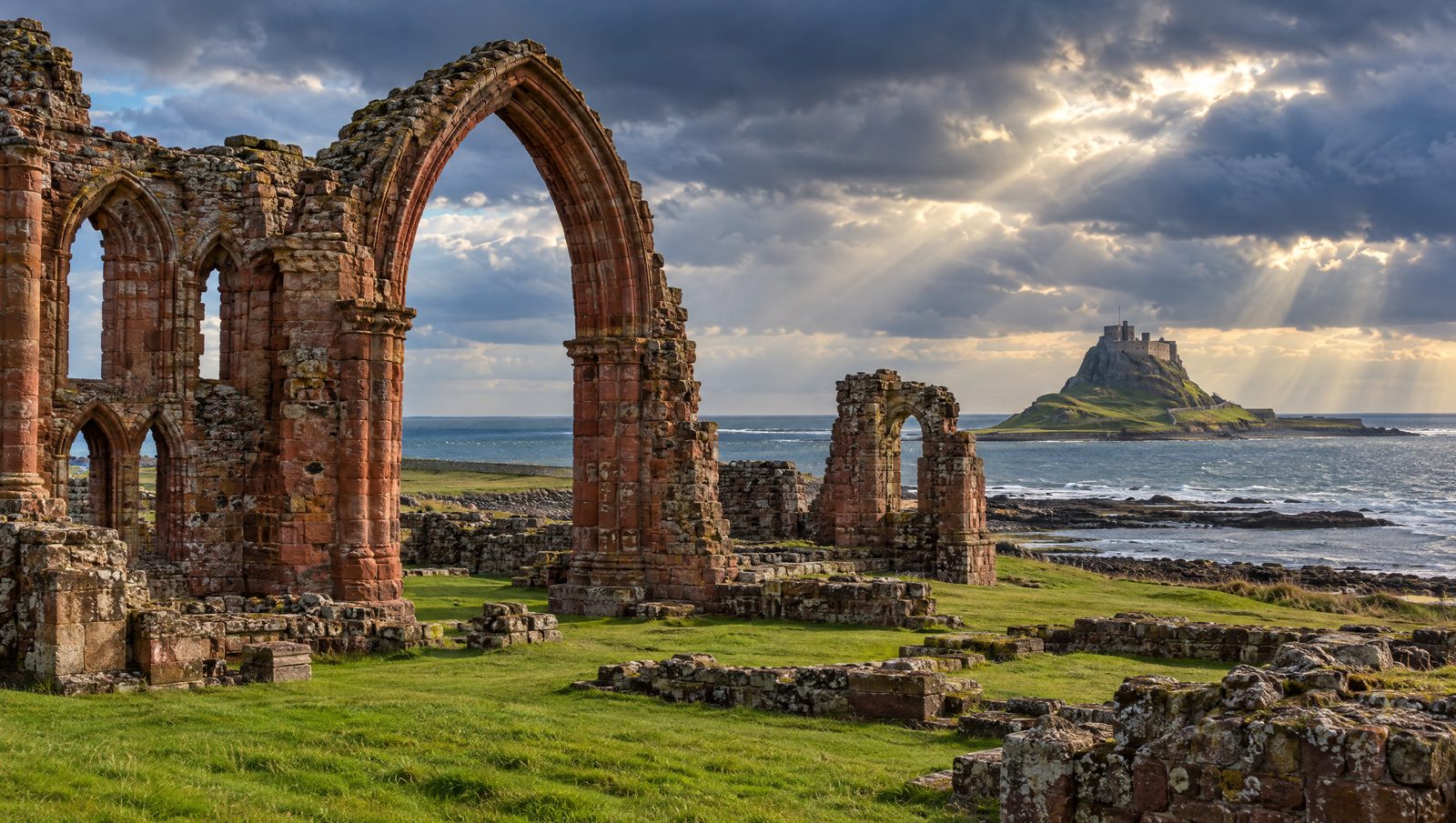 The red sandstone arches of Lindisfarne Priory ruins on Holy Island with Lindisfarne Castle across the bay under a stormy sky