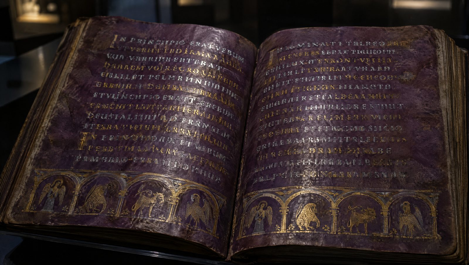 An open manuscript of purple-dyed parchment with lines of Gothic script in silver and gold ink and arcaded decoration at the foot