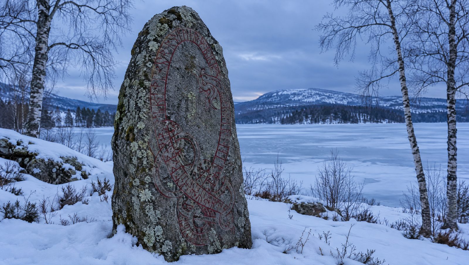 A grey granite runestone carved with a band of runes and knotwork standing in snow beside a frozen lake